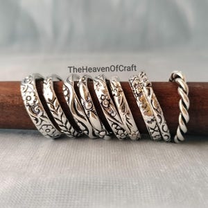 May include: A collection of eight silver-toned metal rings arranged in a row on a dark brown wooden rod. The rings feature various embossed patterns including floral motifs, leaves, wavy lines, and a twisted rope design. The text TheHeavenOfCraft is visible.
