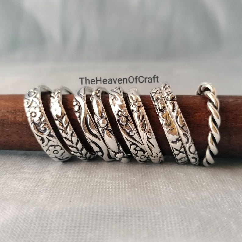 May include: A collection of eight silver-toned metal rings arranged in a row on a dark brown wooden rod. The rings feature various embossed patterns including floral motifs, leaves, wavy lines, and a twisted rope design. The text TheHeavenOfCraft is visible.