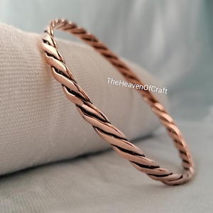 Op de afbeelding: Een gepolijste koperen armband met een gedraaid touwontwerp. Het metaal heeft een warme, metaalachtige roodbruine tint met een gladde, reflecterende afwerking. De tekst TheHeavenOfCraft is zichtbaar in witte letters op de armband.