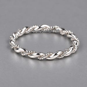 Puede incluir: Un anillo de plata pulida con un diseño de cuerda trenzada que alterna entre segmentos lisos y redondeados y secciones de alambre trenzado con textura fina. El texto TheHeavenOfCraft está impreso en negro en el centro del anillo.