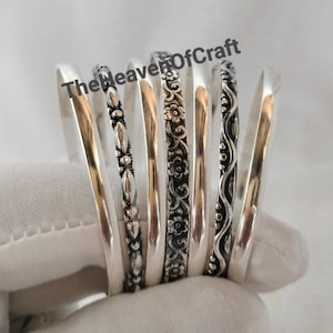 Könnte beinhalten: Ein Set aus sechs silberfarbenen Metallarmreifen mit verschiedenen Designs, darunter glatte Oberflächen, Blumenmuster, Perlenstrukturen und Wellenlinien. Der Text TheHeavenOfCraft ist oben auf den Armreifen zu sehen.