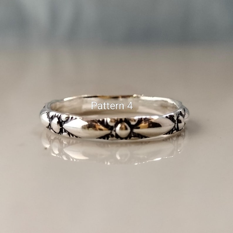May include: A silver-toned metal ring featuring a repeating embossed pattern of alternating elongated leaf-like shapes and small circular dots. The text Pattern 4 is printed in white across the centre of the ring band, which has a polished finish.