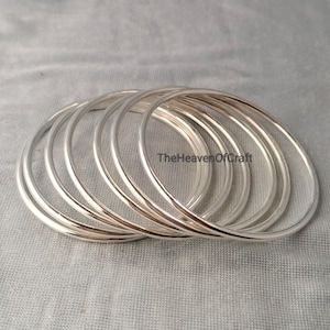 Op de afbeelding: Een set van zeven gladde, gepolijste zilverkleurige armbanden die in een licht overlappende stapel zijn gerangschikt. De tekst TheHeavenOfCraft is duidelijk zichtbaar op het oppervlak van de middelste armband, met een eenvoudig, rond ontwerp.