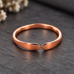 May include: A rose gold-toned open ring with a smooth, polished finish. The inner band features the engraved text TheHeavenOfCraft. The ring has a simple, circular design with two rounded, separated ends that do not meet in the centre.
