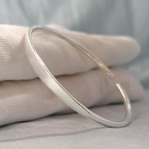 Op de afbeelding: Een zilverkleurige open armband met een gladde, gepolijste afwerking. De armband heeft een eenvoudig, cirkelvormig ontwerp met een opening in het midden en toont een reflecterend metalen oppervlak dat het licht opvangt langs de slanke, gebogen band.