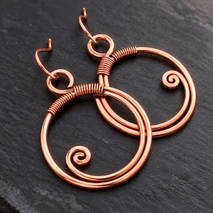 Puede incluir: Un par de pendientes de alambre de cobre con un diseño circular y un extremo en espiral. Cada pendiente está formado por dos aros de alambre concéntricos unidos por segmentos de alambre enrollado y cuenta con un gancho curvado.
