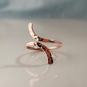 May include: A rose gold-toned bypass ring featuring a hammered, textured finish. The ring has an open design with two tapered ends that curve in opposite directions, creating a wave-like silhouette with a polished, reflective metallic surface.