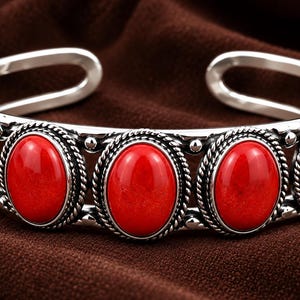 Peut inclure: Un bracelet manchette en argent orné de cinq pierres ovales d'un rouge éclatant, serties individuellement. La bande métallique présente une texture en corde torsadée autour de chaque pierre, avec de petites sphères d'argent entre elles.