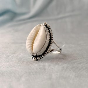 Peut inclure: Une bague en argent avec un coquillage cauri blanc central serti dans une bordure en métal perlé. L'anneau est en métal argenté poli. Le texte TheHeavenOfCraft est visible sur le côté de la bague, qui présente une finition métallique lisse.