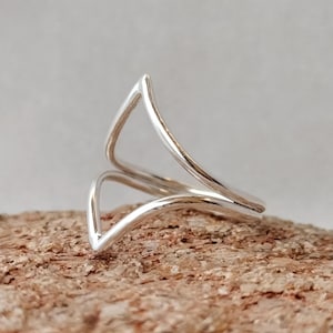 Könnte beinhalten: Ein Ring aus poliertem Silber mit einem zweilagigen Chevron- oder V-förmigen Design. Das Band besteht aus zwei separaten, parallelen Silberdrähten, die an einer scharfen Spitze zusammenlaufen und eine offene, geometrische Struktur bilden.