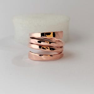Puede incluir: Un anillo de tono oro rosa con un diseño en espiral de varias capas. La banda tiene una superficie pulida y reflectante con una textura orgánica y ondulada que se envuelve, creando un efecto de bobina continua con acabado metálico.