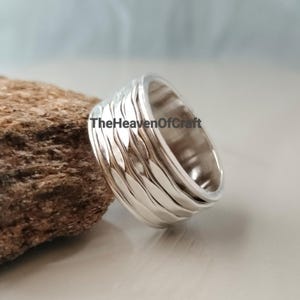 Puede incluir: Un anillo ancho de plata con una superficie texturizada, ondulada y martillada. La banda tiene un acabado pulido y reflectante e incluye el texto TheHeavenOfCraft impreso claramente en la superficie exterior de la banda de metal.