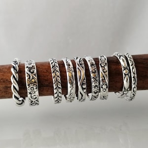 Puede incluir: Una colección de nueve anillos de tono plateado dispuestos en fila sobre una varilla de madera oscura. Los anillos presentan varios patrones en relieve, incluyendo cuerda trenzada, motivos florales, diseños de hojas, líneas onduladas y corazones.