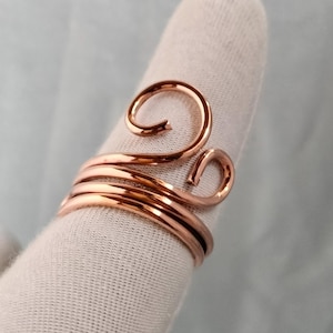 Può includere: Un anello in filo color rame con un design avvolgente a più strati. Il filo metallico è avvolto attorno al dito e termina con due distinti anelli a spirale arrotondati sulla parte superiore. La superficie del metallo è liscia e lucida.