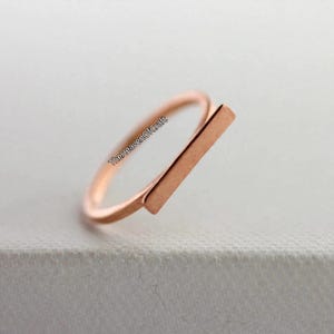 May include: A rose gold-toned ring featuring a thin, circular band and a flat, rectangular bar element positioned horizontally across the top. The text TheHeavenOfCraft is printed in black along the side of the ring band.