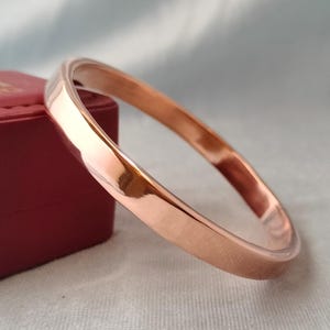 Może przedstawiać: Polerowana bransoletka typu bangle w odcieniu różowego złota o gładkiej, odblaskowej powierzchni i prostym, ciągłym, okrągłym kształcie. Metal ma ciepły, metaliczny połysk, który odbija światło, prezentując czysty, jednolity i pozbawiony zdobień wygląd.