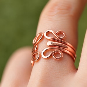 Peut inclure: Une bague en fil de couleur cuivre présentant un design ouvert avec des extrémités en forme de boucles tourbillonnantes. La surface métallique est lisse et polie, affichant un ton chaud. Le texte TheHeavenOfCraft est visible sur l'anneau.