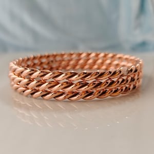 May include: A set of three stacked bangles featuring a polished copper-toned finish. Each bangle is crafted with a continuous twisted rope design, creating a textured, metallic appearance that reflects light across the rounded, spiraling surfaces.