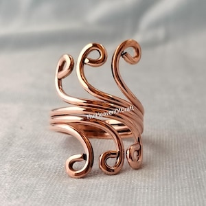 Può includere: Un anello in filo di rame caratterizzato da una serie di linee curve e vorticose che formano un design aperto e stratificato. Il metallo ha una finitura lucida e riflettente. Il testo TheHeavenOfCraft è visibile sulla struttura del filo.