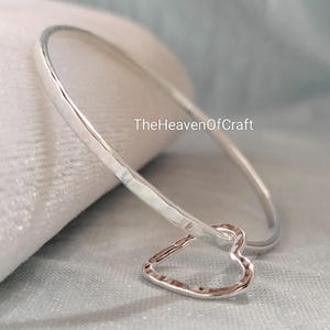 May include: A silver-coloured bangle bracelet featuring a hammered texture and a single dangling heart-shaped charm. The text TheHeavenOfCraft is visible in the centre of the image. The bracelet has a smooth, reflective finish and a circular shape.