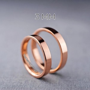 May include: Two rose gold-coloured metal rings with a smooth, flat band design. The rings are positioned one behind the other. The text 3 MM is printed above the rings, and the text TheHeavenOfCraft is printed on the side of the front ring.