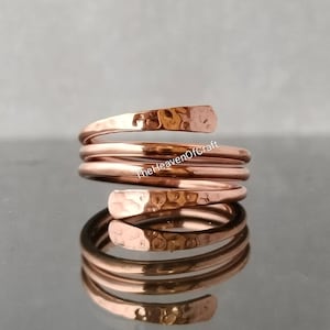 Peut inclure: Une bague en cuivre présentant un design en spirale avec trois bobines distinctes. La surface métallique a une texture martelée aux extrémités, et le texte TheHeavenOfCraft est visible au centre de la bague, se reflétant sur la surface.