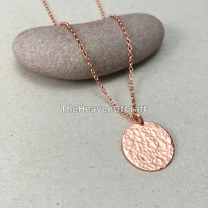 Pode incluir: Um colar em tom de ouro rosa com uma corrente delicada e um pingente redondo de textura martelada. A superfície metálica do disco reflete a luz com um padrão de covinhas, suspenso por uma corrente de elos finos sobre uma pedra lisa.