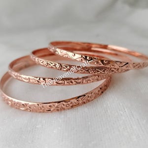 Peut inclure: Un ensemble de quatre bracelets rigides en métal cuivré, chacun présentant un motif floral ou géométrique en relief distinct sur la surface extérieure. Les bracelets sont empilés et se chevauchent, avec le texte TheHeavenOfCraft visible.
