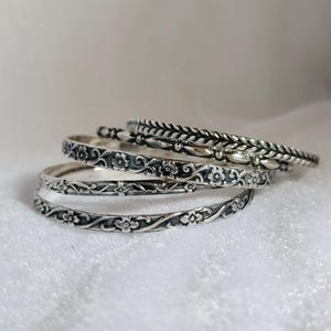 May include: A set of four silver-toned metal bangles stacked together. Each bangle features a distinct embossed pattern, including floral motifs, scrolling vines, and a braided texture, all with darkened recessed areas to highlight the details.