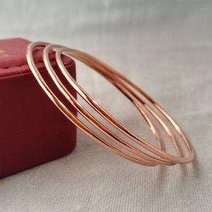 May include: A set of three thin, polished rose gold-colored metal bangles. The circular bracelets feature a smooth, reflective surface and a simple, continuous design. These metallic bangles are stacked together, showcasing their warm, metallic hue.