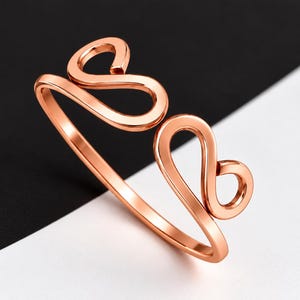 Pode incluir: Um anel aberto em tom de ouro rosa com dois motivos simétricos em forma de laço em cada extremidade da banda. O metal tem um acabamento liso e polido, e o design é caracterizado pelas suas linhas contínuas, fluidas e curvas.