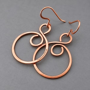 Puede incluir: Un par de pendientes colgantes de cobre con un diseño de alambre circular y un bucle de infinito en la parte superior. El metal tiene un acabado cálido. El texto TheHeavenOfCraft es visible en la curva inferior de uno de los pendientes.