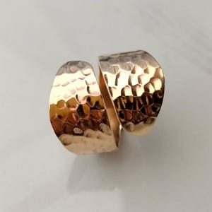 Puede incluir: Un anillo de tono dorado con un diseño de banda ancha abierta. La superficie del metal presenta una textura martillada distintiva, creando una serie de pequeñas hendiduras reflectantes en todo el exterior de esta pieza de joyería.