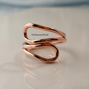Pode incluir: Um anel em tom de ouro rosa com um design aberto estilo bypass, apresentando dois laços fluidos em forma de gota que se sobrepõem. A superfície metálica é lisa e reflexiva. O texto TheHeavenOfCraft é visível no centro da estrutura do anel.