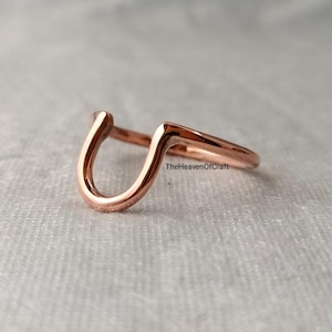 May include: A rose gold ring featuring a distinct U-shaped or chevron-style contour. The metal surface is smooth and polished, reflecting light. The text TheHeavenOfCraft is clearly visible on the side of the band, indicating the brand name.
