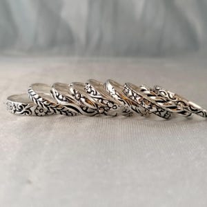 May include: A collection of seven silver-toned metal rings arranged in a row, each featuring distinct embossed patterns including floral motifs, twisted rope textures, and intricate scrollwork designs with oxidised detailing in the recessed areas.