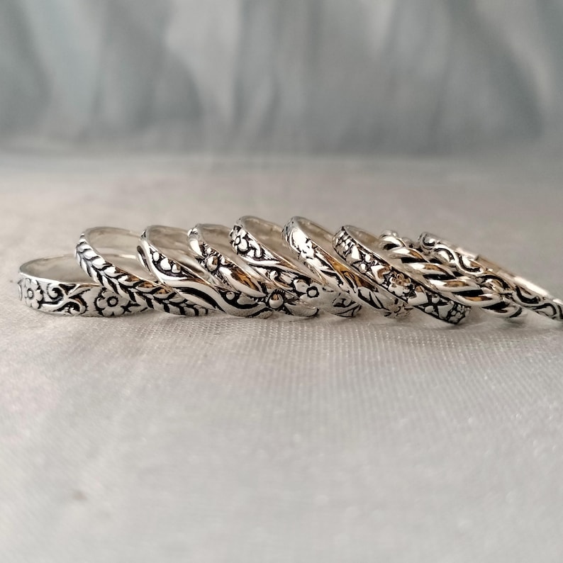 May include: A collection of seven silver-toned metal rings arranged in a row, each featuring distinct embossed patterns including floral motifs, twisted rope textures, and intricate scrollwork designs with oxidised detailing in the recessed areas.