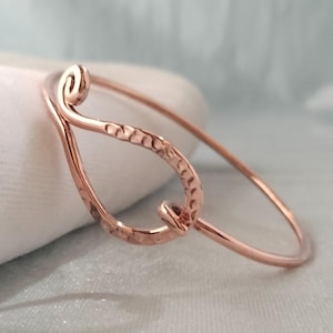 May include: A rose gold-toned metal bangle bracelet featuring a teardrop-shaped focal point. The wire is hammered to create a textured surface, and the ends of the wire are curled into small, tight spirals at the top of the teardrop design.