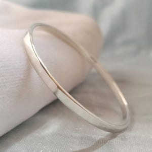 May include: A polished silver bangle bracelet with a smooth, rounded exterior surface and a reflective metallic finish. The circular band features a consistent thickness throughout its design, showcasing a clean and simple aesthetic with no additional embellishments.