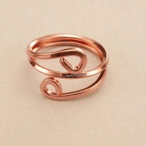 Op de afbeelding: Een koperkleurige metalen ring met een bypass-ontwerp met twee open, geluste uiteinden. De band is gemaakt van een enkele doorlopende draad die rondloopt om de cirkelvorm te vormen. De tekst TheHeavenOfCraft is zichtbaar op de ringband.