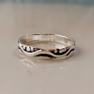 May include: A silver ring featuring a continuous embossed wave pattern with darkened recessed areas and the text Pattern 3 printed in white. The band has a polished metallic finish and a rounded profile, showcasing a detailed design throughout.