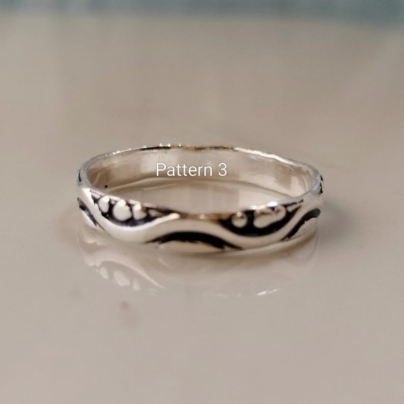 May include: A silver ring featuring a continuous embossed wave pattern with darkened recessed areas and the text Pattern 3 printed in white. The band has a polished metallic finish and a rounded profile, showcasing a detailed design throughout.