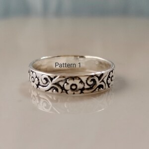 May include: A silver-toned metal ring featuring a continuous embossed floral and scrollwork pattern around the exterior band. The interior of the band is smooth and clearly displays the text Pattern 1 printed in a dark grey font.