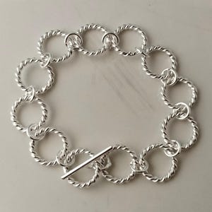 May include: A silver-toned bracelet composed of a series of interconnected circular links, each featuring a twisted rope-like texture. The bracelet is secured with a T-bar toggle clasp, which consists of a straight metal bar that passes through a ring.
