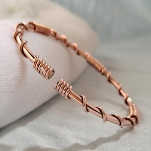 Può includere: Un bracciale a fascia in filo metallico color oro rosa, composto da un cerchio principale avvolto da spirali di filo più sottile a intervalli regolari, con due sezioni cilindriche avvolte strettamente alle estremità aperte.