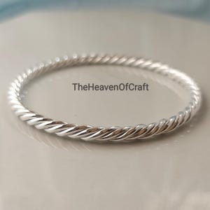 May include: A polished silver-toned bangle bracelet featuring a continuous twisted rope texture throughout the entire band. The text TheHeavenOfCraft is printed in black in the center of the circular opening of the metallic jewelry piece.
