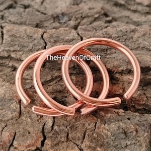 Puede incluir: Tres anillos circulares de cobre con un acabado liso y pulido. Los anillos están abiertos, formando una figura en C. El texto TheHeavenOfCraft está impreso en negro sobre el centro de los anillos, dispuestos en un patrón superpuesto.
