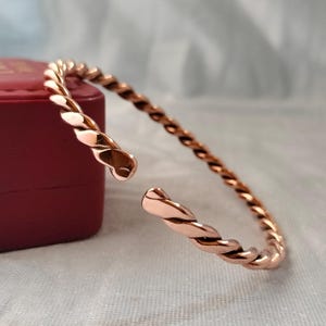 Könnte beinhalten: Ein Armreif in Roségold-Optik mit einem durchgehenden gedrehten Seildesign. Der Armreif ist offen gestaltet und endet an der Öffnung in zwei abgerundeten, glatten Endstücken. Die Metalloberfläche hat ein poliertes, reflektierendes Finish.