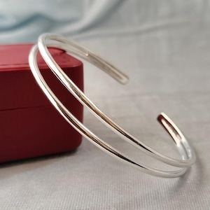 Peut inclure: Un bracelet manchette argenté poli présentant un design à double anneau avec une surface lisse et réfléchissante. Ce bracelet ouvert a une forme circulaire et est positionné devant une petite boîte à bijoux rectangulaire de couleur rouge.