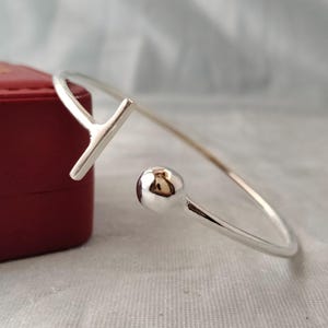Könnte beinhalten: Ein silberfarbenes, offenes Armband mit glatter, polierter Oberfläche. Ein Ende des Armbands endet in einer kleinen, abgerundeten Kugel, während das andere Ende ein T-förmiges Stangendesign aufweist. Das Metall hat eine reflektierende Oberfläche.
